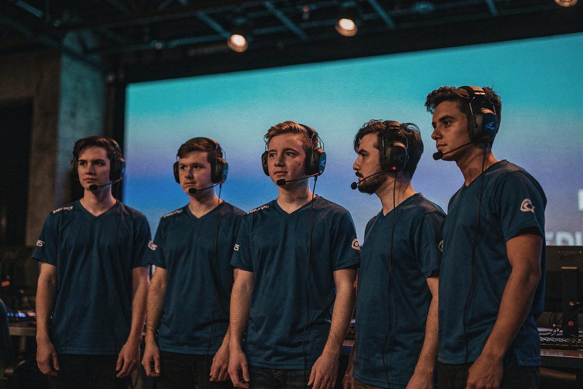 Five players in matching jerseys and headsets standing in a dim arena-style space for a roster photograph